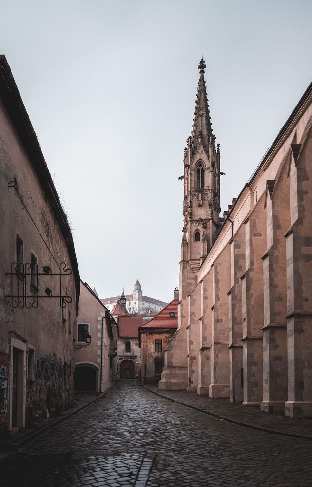 Un callejón estrecho de adoquines entre edificios antiguos, que conduce hacia una alta aguja gótica con un castillo en una colina a lo lejos.