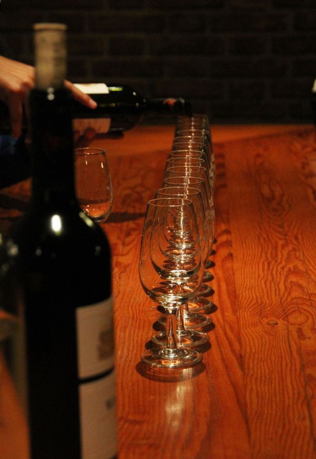 Una mano vierte vino tinto de una botella en una larga fila de copas vacías sobre una mesa de madera para una cata de vinos.