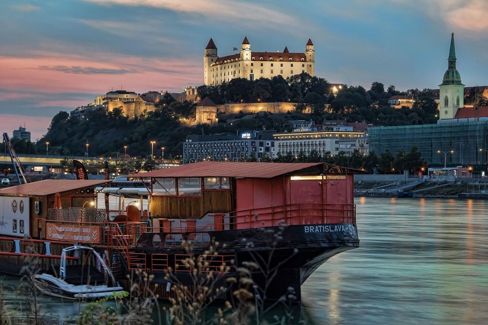 Un gran barco está amarrado en un río con un castillo iluminado y un paisaje urbano en una colina al fondo al anochecer.