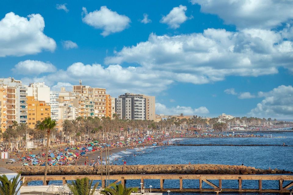 Una vista amplia de una concurrida playa urbana en un día soleado, con gente nadando y relajándose bajo sombrillas coloridas cerca de edificios y palmeras.
