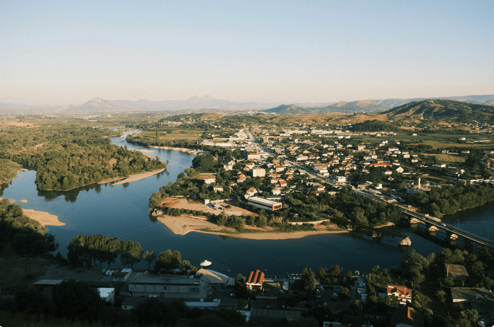 An aerial view of a river curving around a town, with a bridge, green hills, and distant mountains under a clear sky.