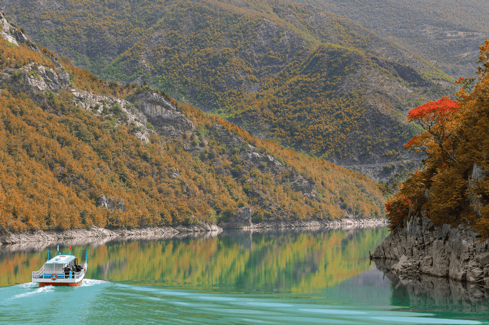 A tour boat carrying a WeRoad group trip navigates a calm lake, surrounded by steep mountains covered in autumn foliage.
