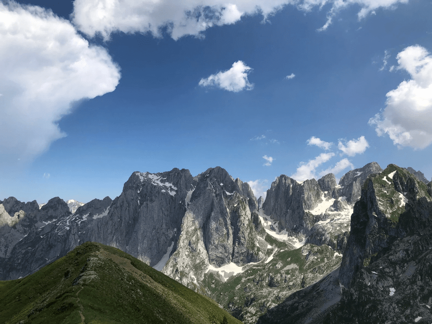 A green grassy ridge leads towards a jagged mountain range with patches of snow under a blue sky with white clouds.
