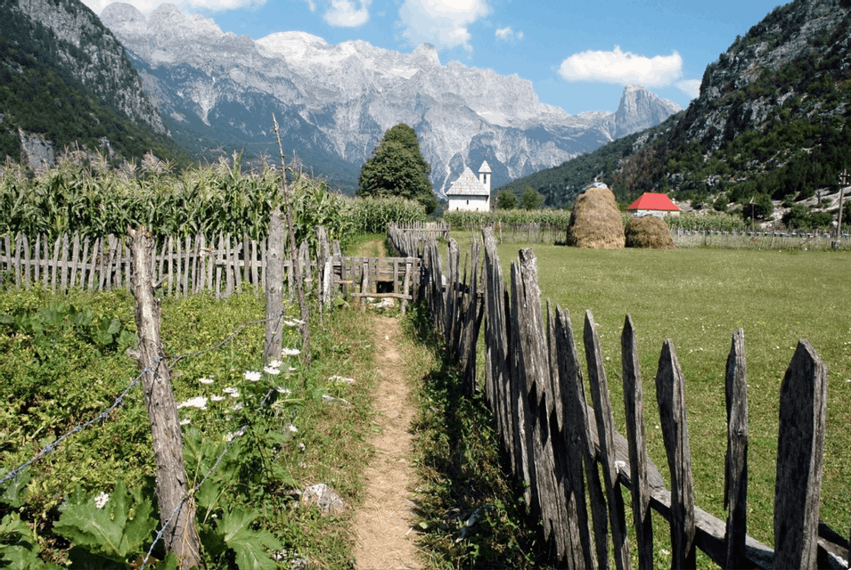 A dirt path along a wooden fence leads through a green valley with a church, cornfield, and tall mountains in the background.