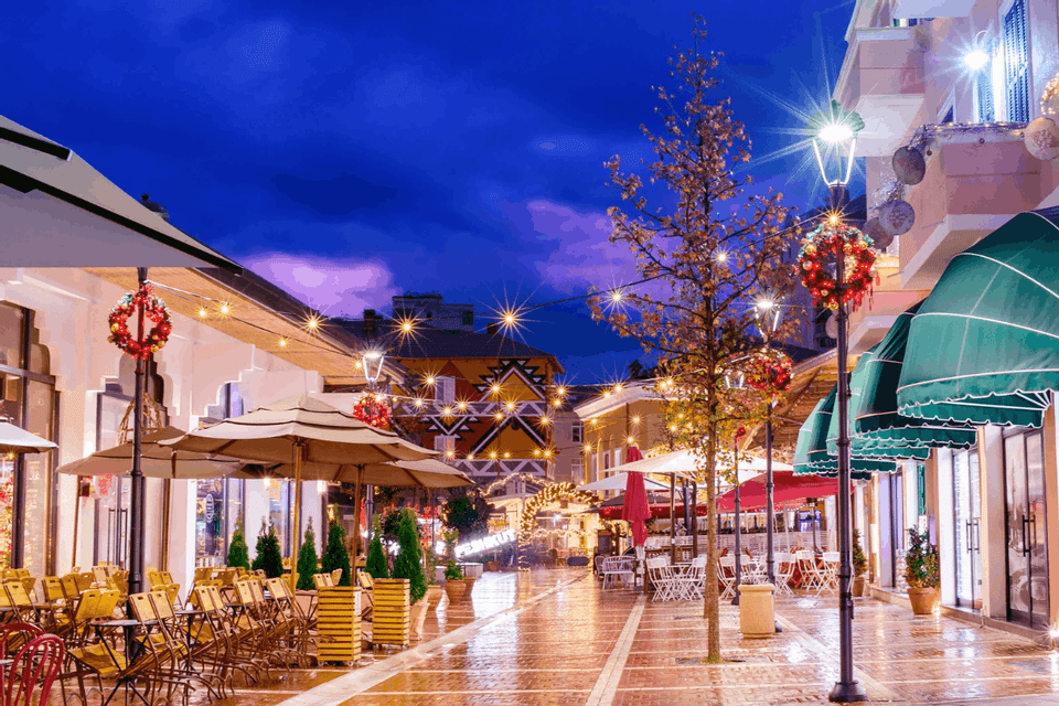 A wet pedestrian street lined with outdoor cafes is illuminated by string lights and decorated with Christmas wreaths at twilight.
