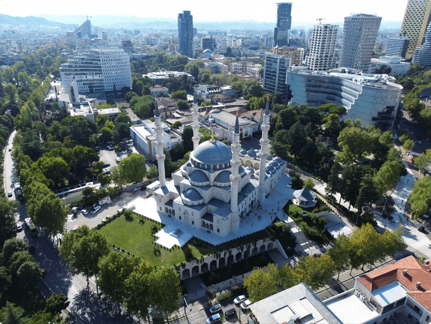 An aerial view of a large mosque with blue domes and four minarets, surrounded by green trees and a modern cityscape.