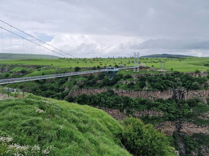 Un pont suspendu en verre avec une structure en forme de diamant au centre enjambe un canyon profond et verdoyant sous un ciel nuageux.