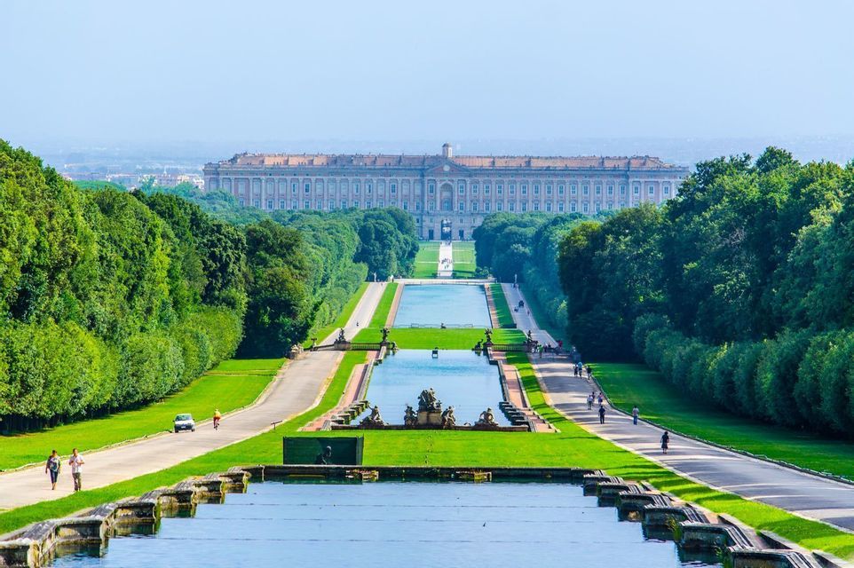 Una vista elevada de un largo jardín formal con fuentes de agua en cascada y caminos arbolados que conducen a un gran palacio.