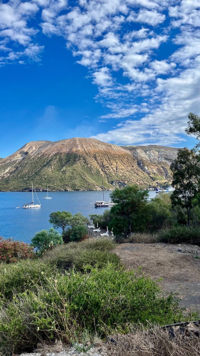 Barche a vela galleggiano in una baia calma ai piedi di una grande isola montuosa e rocciosa sotto un cielo azzurro con nuvole sparse.