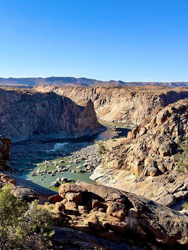Un fiume si snoda attraverso un vasto canyon roccioso con ripide scogliere sotto un cielo azzurro e limpido.