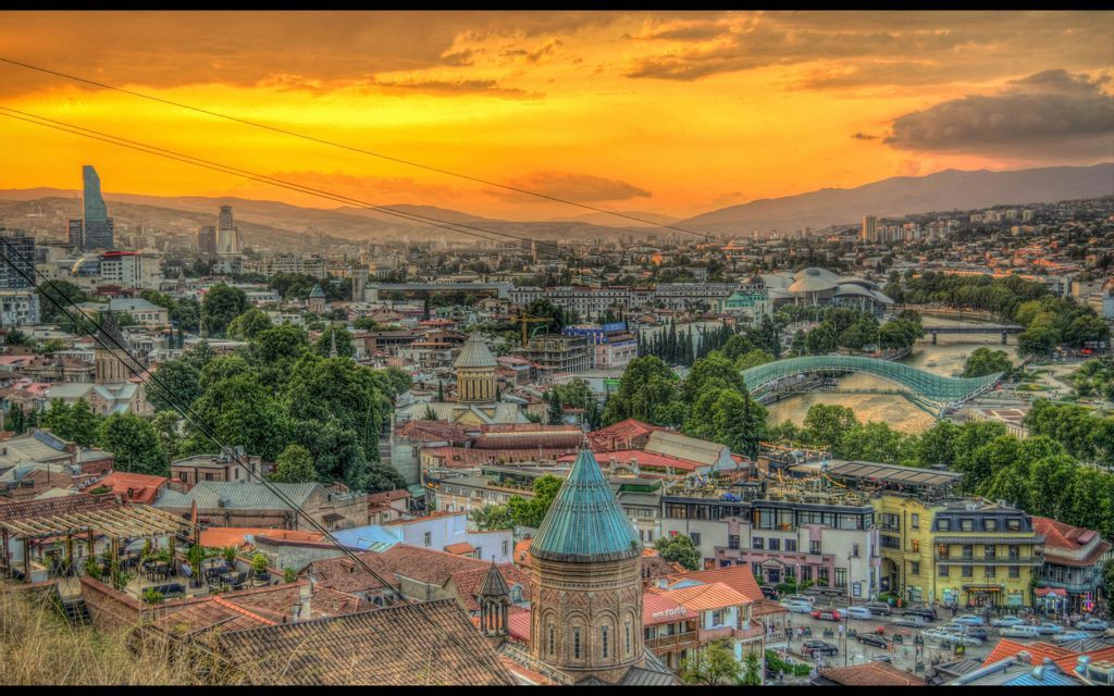 Une vue en hauteur d'une ville avec des bâtiments historiques et modernes, un fleuve et des montagnes sous un coucher de soleil doré.
