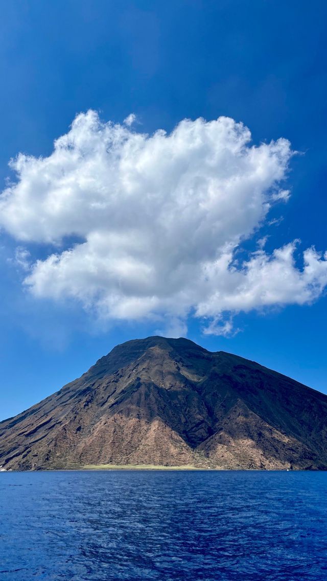 Un grande monte vulcanico sorge dal mare blu, la sua cima in ombra sotto una singola grande nuvola bianca in un cielo azzurro brillante.