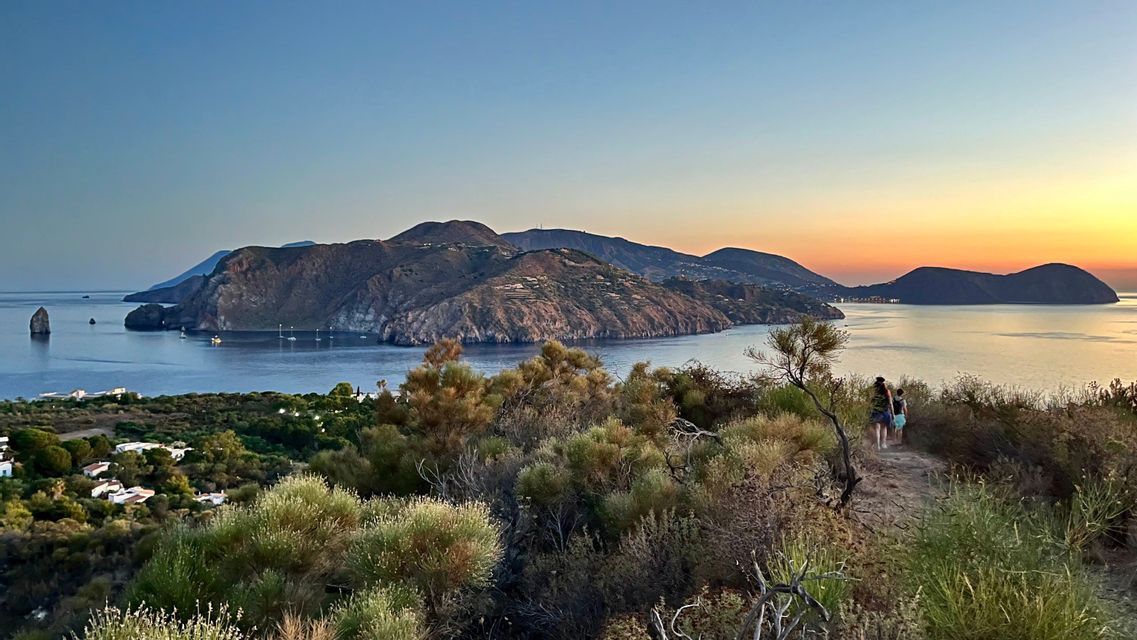 Due persone di un viaggio di gruppo WeRoad camminano su un sentiero collinare che domina una baia con isole vulcaniche al tramonto.