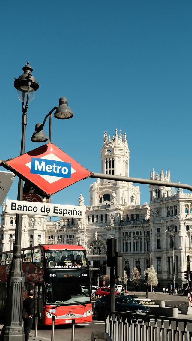 A red Metro sign for the Banco de España station, with a red tour bus and an ornate white building under a clear blue sky.