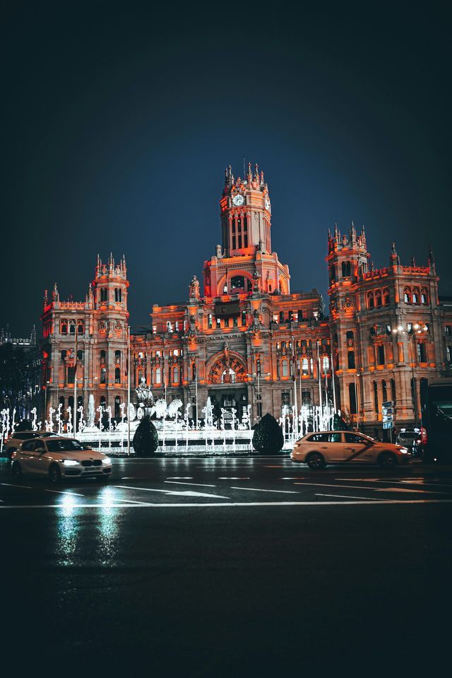 An ornate building with a clock tower illuminated by orange lights at night, with cars driving on a wet street in the foreground.