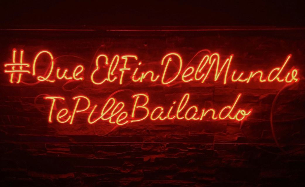 A glowing red neon sign with cursive Spanish text that reads '#QueElFinDelMundo TePilleBailando' on a dark wall.