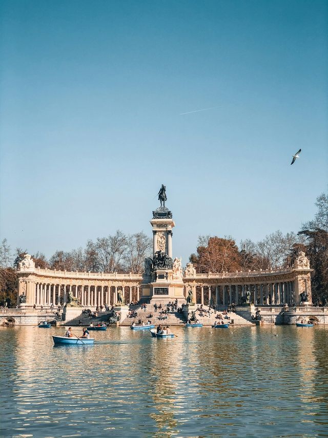 People in blue rowboats paddle on a lake in front of a grand monument featuring an equestrian statue, under a clear blue sky.