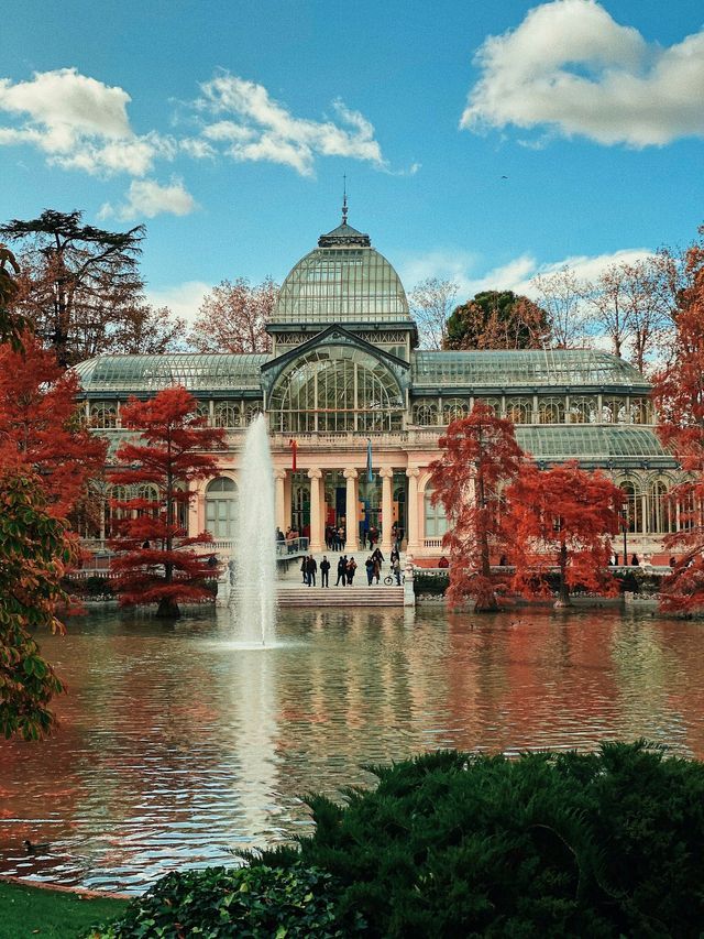 A large glass building sits behind a lake with a central fountain, surrounded by trees with bright red autumn leaves.