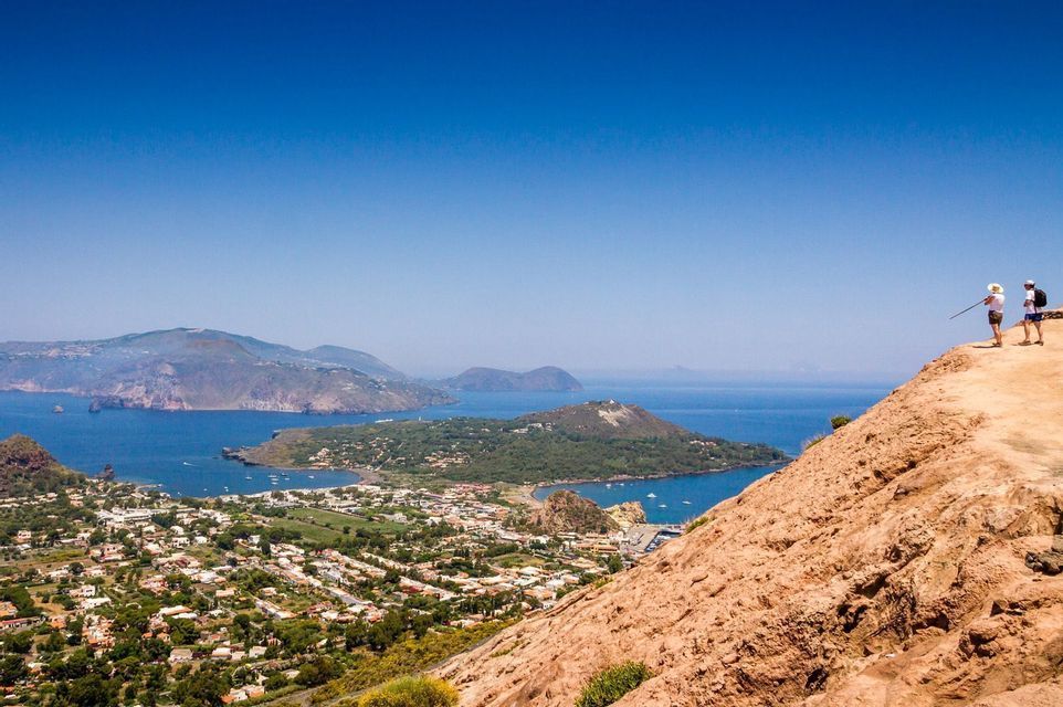 Due partecipanti a un viaggio di gruppo WeRoad si trovano su una scogliera, ammirando una città costiera e diverse isole sotto un cielo limpido e azzurro.