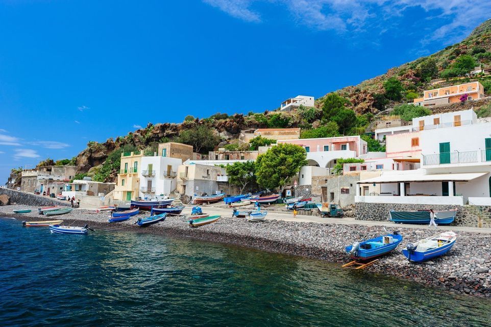 Piccole barche sono ormeggiate su una spiaggia di ciottoli di fronte a un villaggio incastonato su un fianco di collina roccioso, sotto un cielo azzurro.