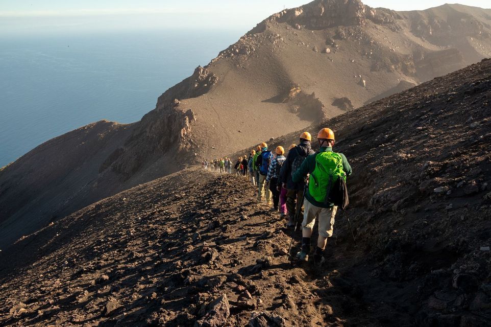 Un gruppo WeRoad con i caschi fa trekking in fila indiana su un ripido sentiero di montagna vulcanico a picco sull'oceano.