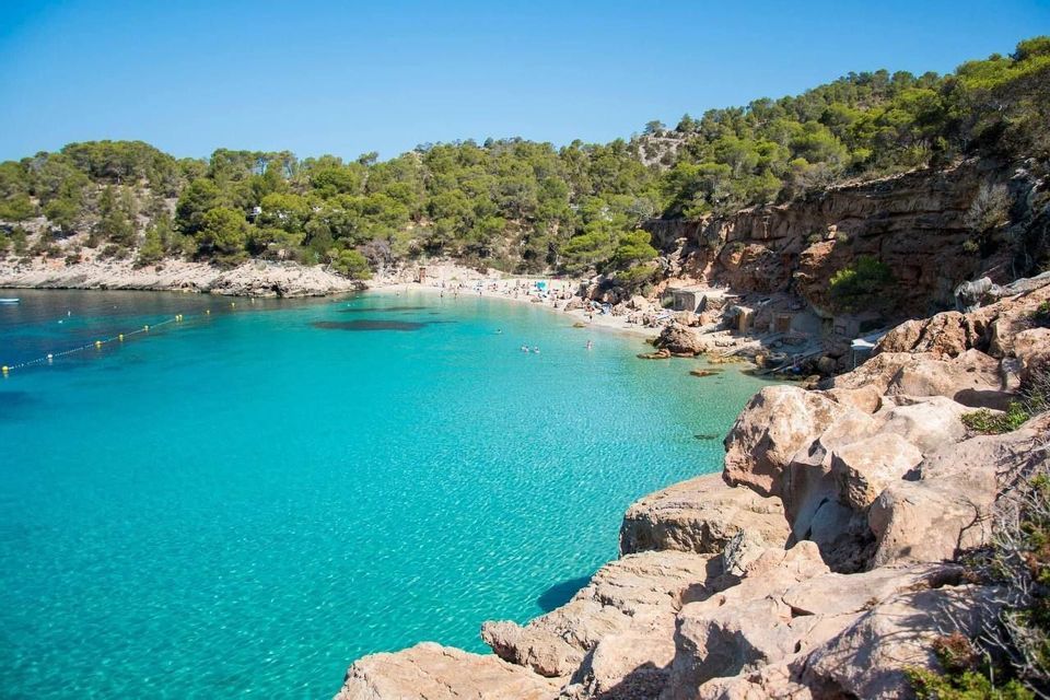 Una vista desde un acantilado rocoso de una cala apartada con agua turquesa cristalina, una playa de arena con gente y una costa arbolada.