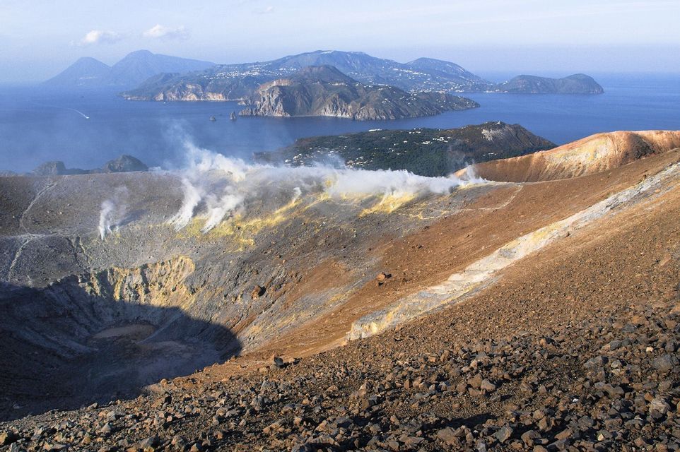 Il vapore sale dai pendii sulfurei di un cratere vulcanico, con vista su isole oltre il mare sullo sfondo.