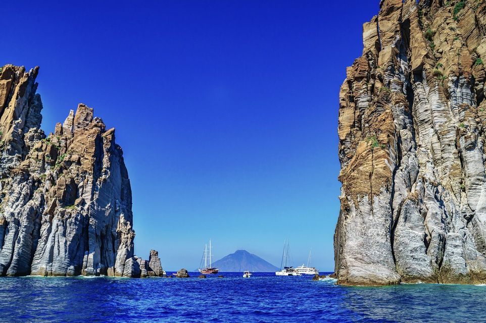 Diverse barche sull'acqua blu passano tra due grandi formazioni rocciose, con un vulcano visibile in lontananza sotto un cielo sereno.