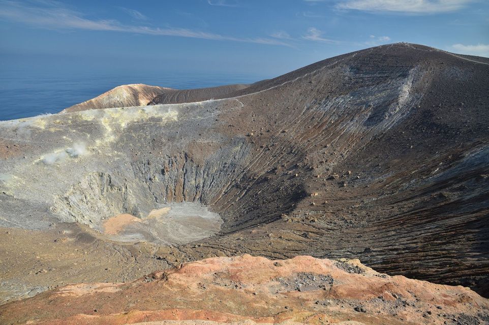 Una vista panoramica di un grande cratere vulcanico con vapore che sale dal suo bordo, con il mare visibile sullo sfondo sotto un cielo blu.