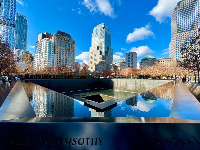 A memorial reflecting pool with engraved names on its parapet, reflecting the surrounding city skyscrapers and a bright blue sky.