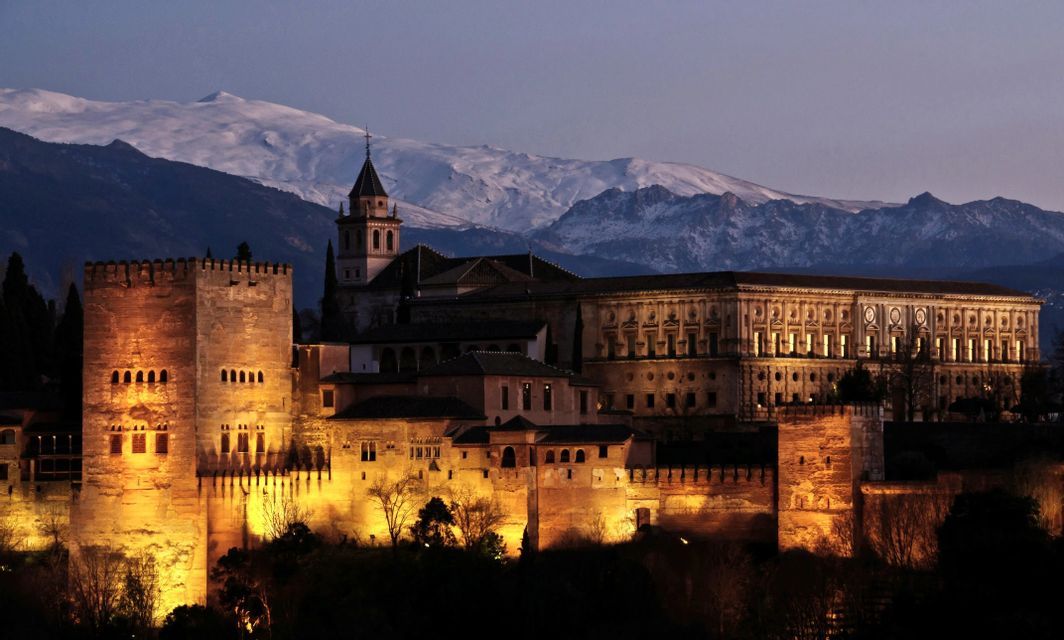 Un gran complejo palaciego de piedra iluminado al anochecer, con montañas cubiertas de nieve a lo lejos.