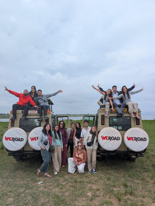 Un grupo de WeRoad posando para una foto frente a dos jeeps de safari en un campo de hierba, con un cuerpo de agua al fondo.