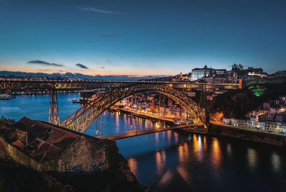 Un ponte ad arco metallico illuminato attraversa un ampio fiume al tramonto, con le luci della città che si riflettono sull'acqua.