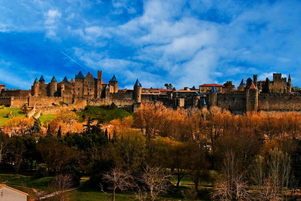 Un château de pierre médiéval avec des tours aux toits bleus se dresse sur une colline au-dessus d'une forêt automnale, sous un ciel bleu éclatant avec des nuages.
