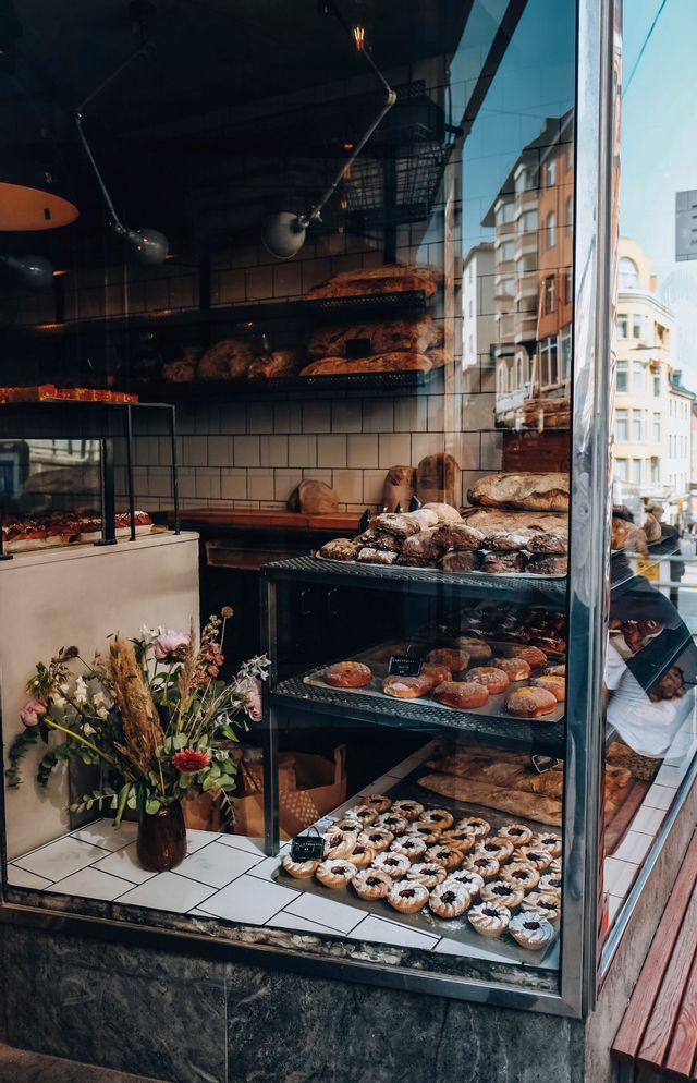 La vitrine d'une boulangerie expose du pain frais, des beignets et des tartelettes à la confiture sur des étagères, avec le reflet d'une rue de la ville dans le verre.