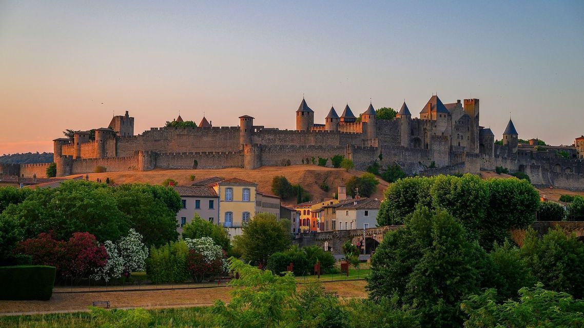 Un grand château de pierre avec de nombreuses tourelles se dresse sur une colline, illuminé par la lumière chaude du coucher de soleil au-dessus d'une petite ville aux arbres verdoyants.