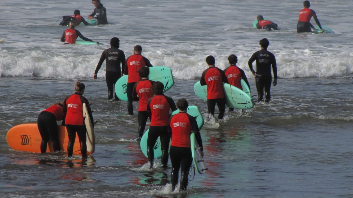 Un groupe WeRoad, en combinaisons de surf, se dirige vers l'océan avec des planches pour une leçon encadrée par leurs moniteurs.