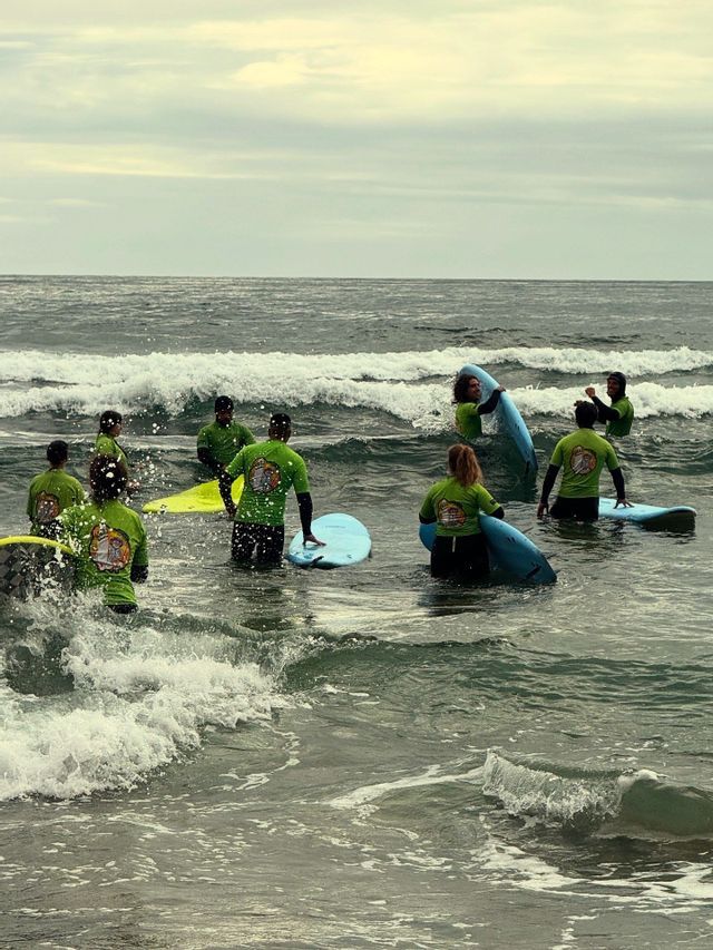 A WeRoad group trip wearing matching green shirts stands with surfboards in the ocean waves during a lesson.