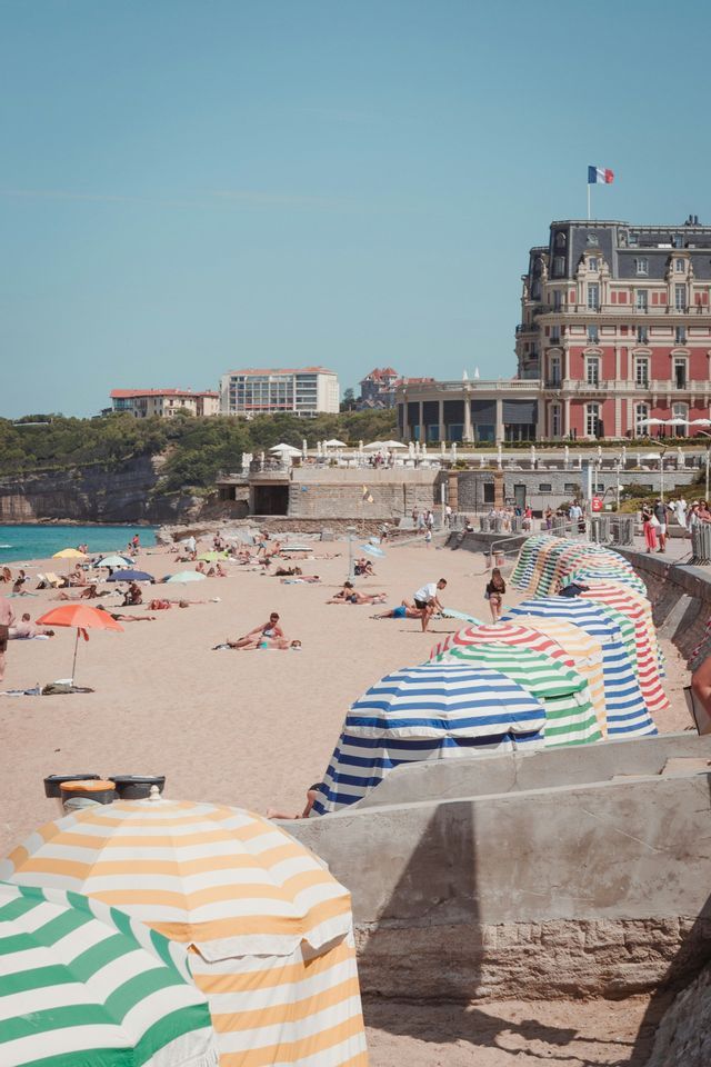 Des gens se prélassent au soleil sur une plage de sable remplie de parasols rayés colorés, devant un grand bâtiment arborant un drapeau français.