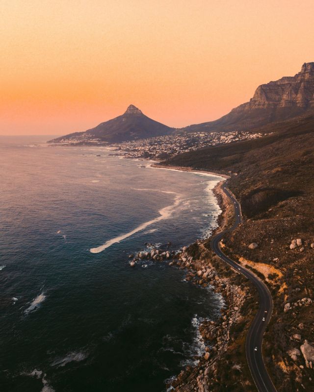 Una vista aerea di una strada tortuosa lungo una costa rocciosa, con montagne e una città in lontananza sotto un cielo arancione al tramonto.