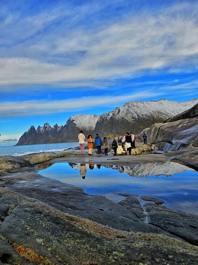 Un viaggio di gruppo WeRoad si trova su una costa rocciosa, con montagne innevate e il cielo riflesso in uno specchio d'acqua.