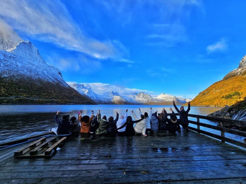 Un grupo de viaje de WeRoad está en un muelle de madera con las manos levantadas, contemplando montañas nevadas al otro lado de un lago bajo un cielo azul.