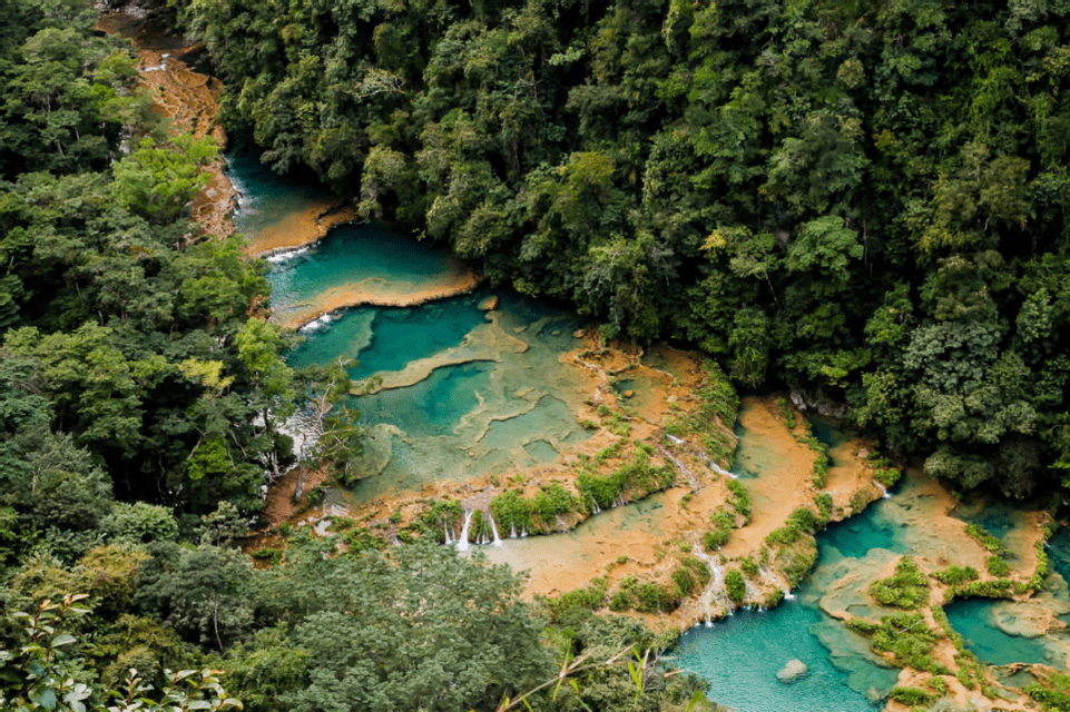 Eine Luftaufnahme eines türkisfarbenen Flusses mit terrassierten Becken und Wasserfällen, der durch einen dichten grünen Wald fließt.