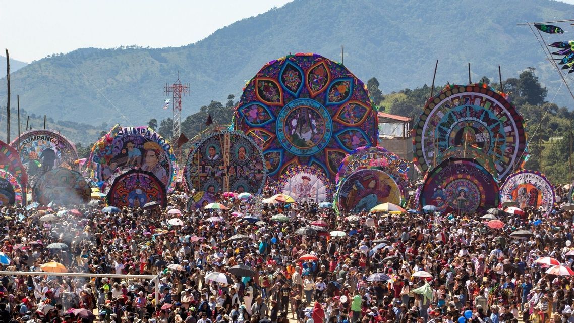 Eine riesige Menschenmenge versammelt sich auf einem Open-Air-Festival, um mehrere riesige, bunte, verzierte Drachen zu bestaunen, die vor einer Bergkette ausgestellt sind.