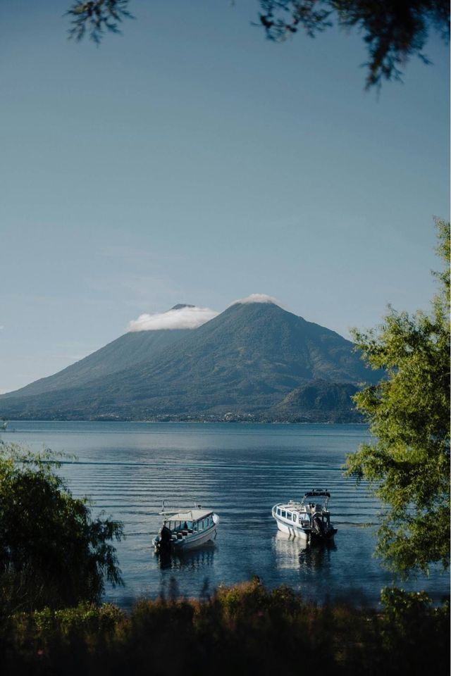 Zwei Motorboote treiben auf einem ruhigen See vor einem großen Berg mit wolkenbedecktem Gipfel, umrahmt von grünem Laub.