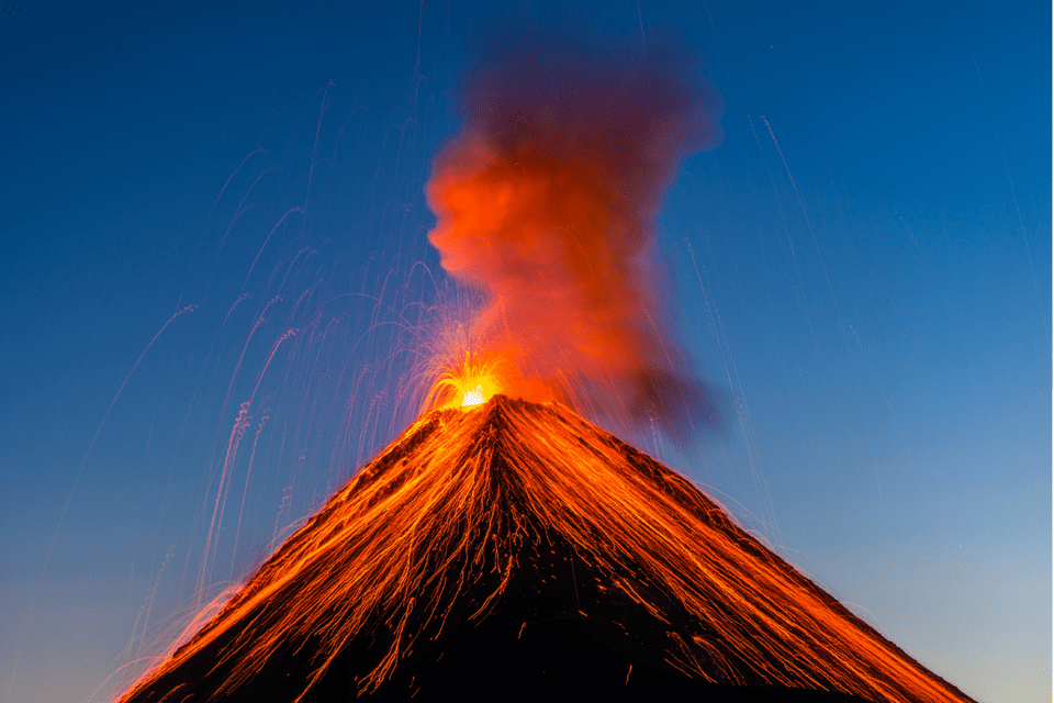 Geschmolzene Lava speit aus dem Gipfel eines ausbrechenden Vulkans und fließt in hellen Strömen dessen Hänge unter blauem Himmel hinab.