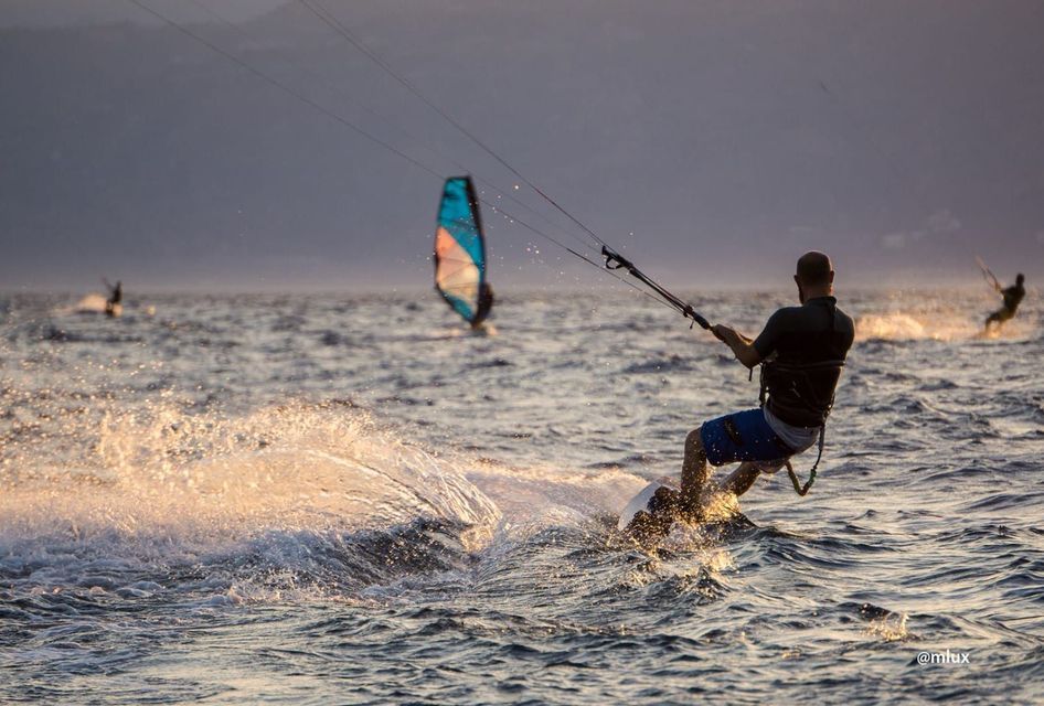Un uomo pratica kitesurf sull'oceano al tramonto, sollevando uno spruzzo. Sullo sfondo, altre persone fanno windsurf e kitesurf.