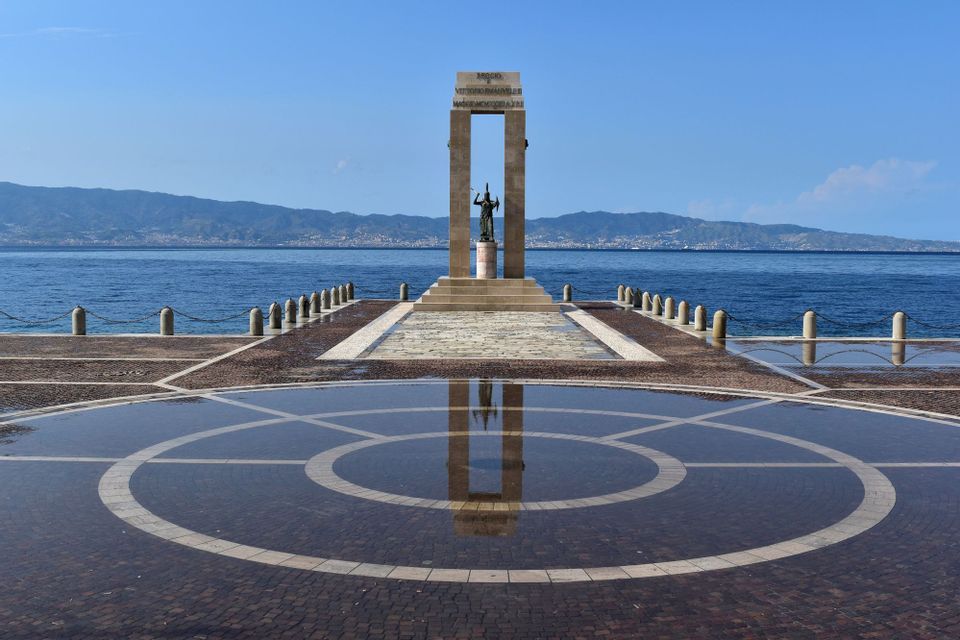 Un monumento su un molo di pietra si riflette in una grande pozzanghera, con il mare e le montagne sullo sfondo.