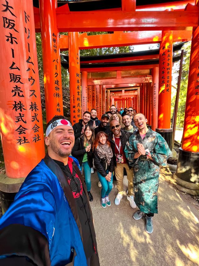 Ein Mann im blauen Gewand macht ein Selfie mit einer WeRoad-Gruppe, die alle unter einem Tunnel aus roten japanischen Torii-Toren lächeln.