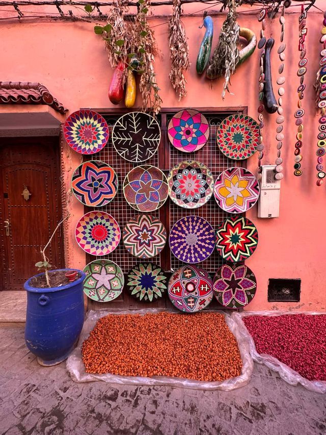 Des assiettes et paniers tressés colorés sont exposés à la vente sur un présentoir devant un mur rose, au-dessus de tas d'épices dans une rue pavée.